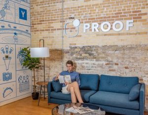 Young person sat on a blue sofa reading a magazine. The word proof is above on the wall